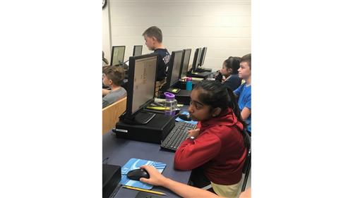 A girl peeks over her computer to look at the board while other students sit behind desk top computers working on Scratch. 