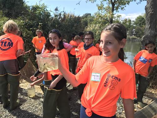Girl holds small aquarium with fish caught while students in the background wearing fishing boots look on. 