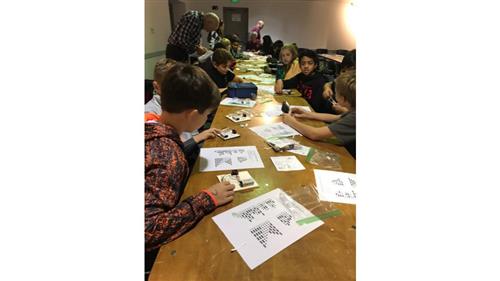 Ten students sitting on each side of a long table work on electromagnetic sensors with a schematic. 